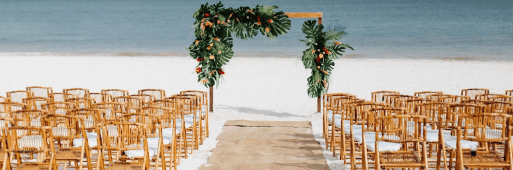 A destination wedding setup with wooden chairs and a rectangular arch with big green fern leaves and flowers with the ocean in the background