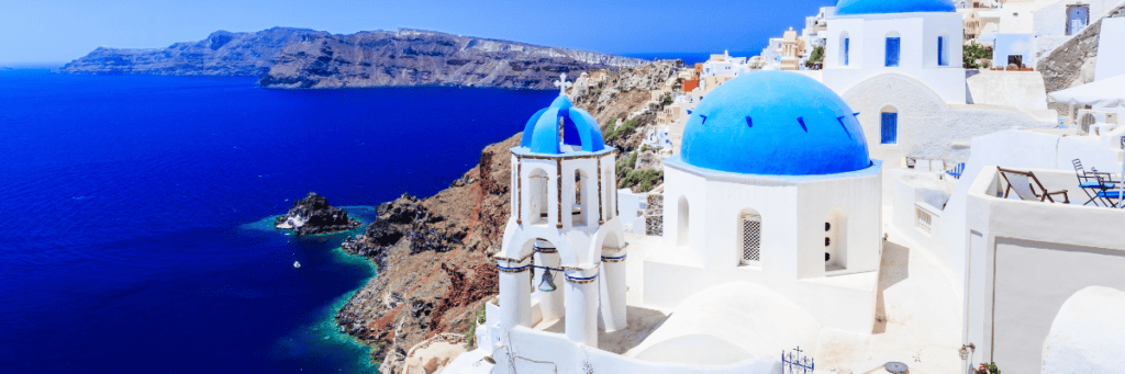 View of white buildings with blue roofs in Greece overlooking the Mediterranean with mountains in the background