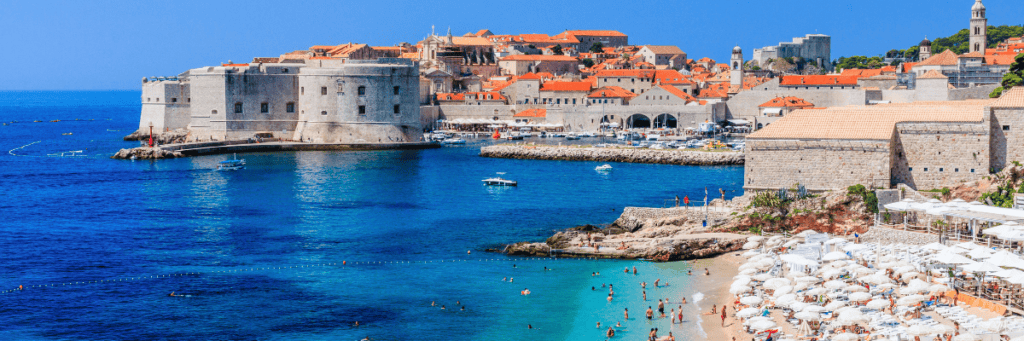 Crystal blue waters in Croatia with buildings with orange roofs along the water line and a beach area with white umbrellas