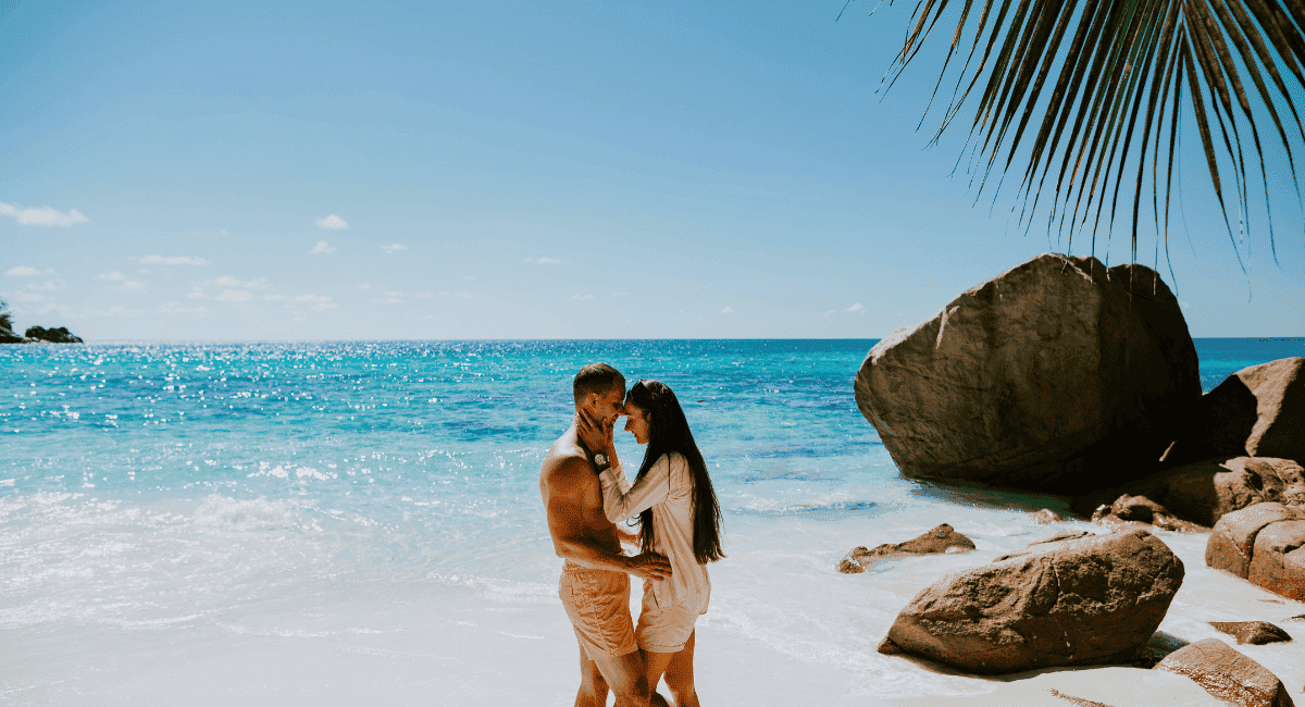 A man and a woman posing with their heads together on the beach with the ocean and some big boulders in the background