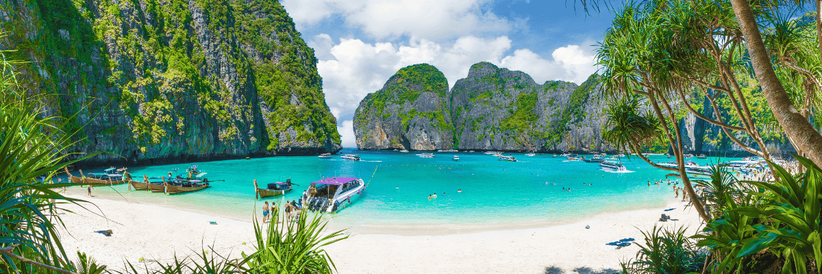 A Thailand beach with people on the sand and boats in the water with the green/rocky cliffs in the distance