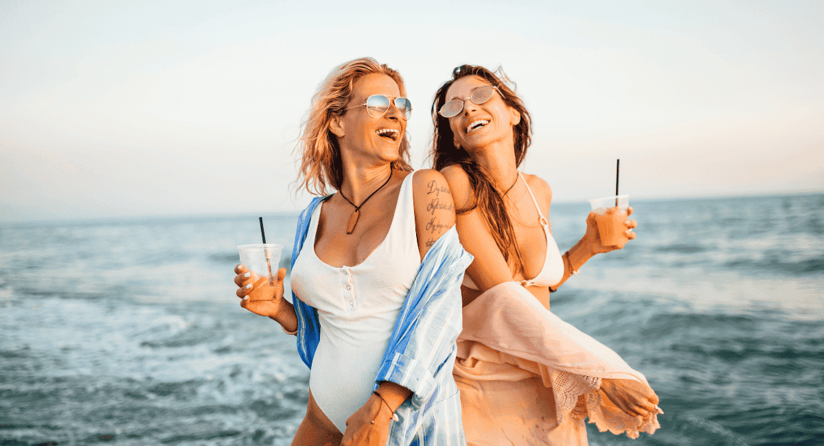 Two women in swimsuits and coverups standing next to each other laughing and drinking a fruity drink on the beach