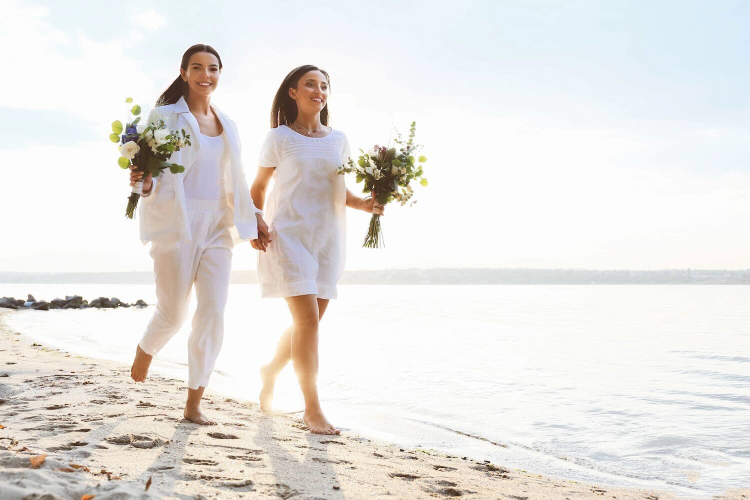 Two women in swimsuits and coverups standing next to each other laughing and drinking a fruity drink on the beach