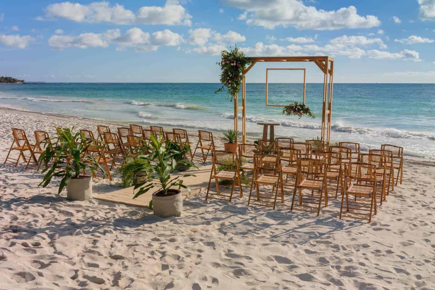 A wooden wedding arch for a destination wedding and rows of chairs are arranged on a sandy beach with plants and the ocean in the background under a partly cloudy sky.