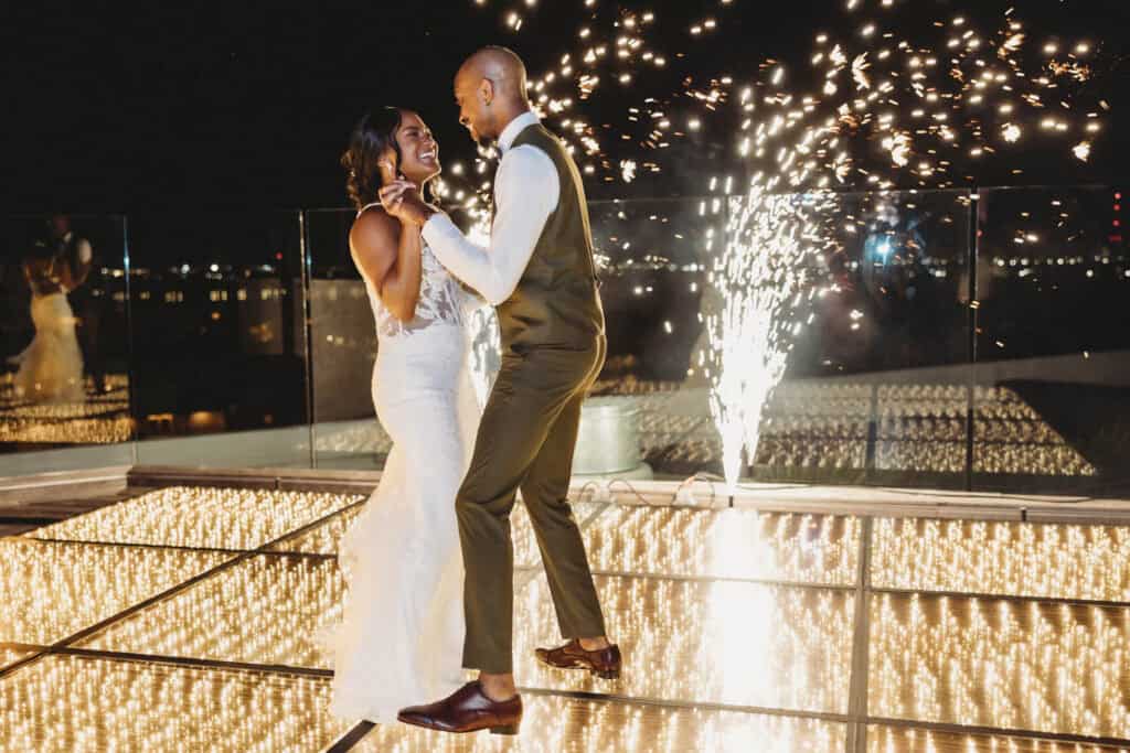 A bride and groom dance together on a lit floor at night with fireworks in the background.