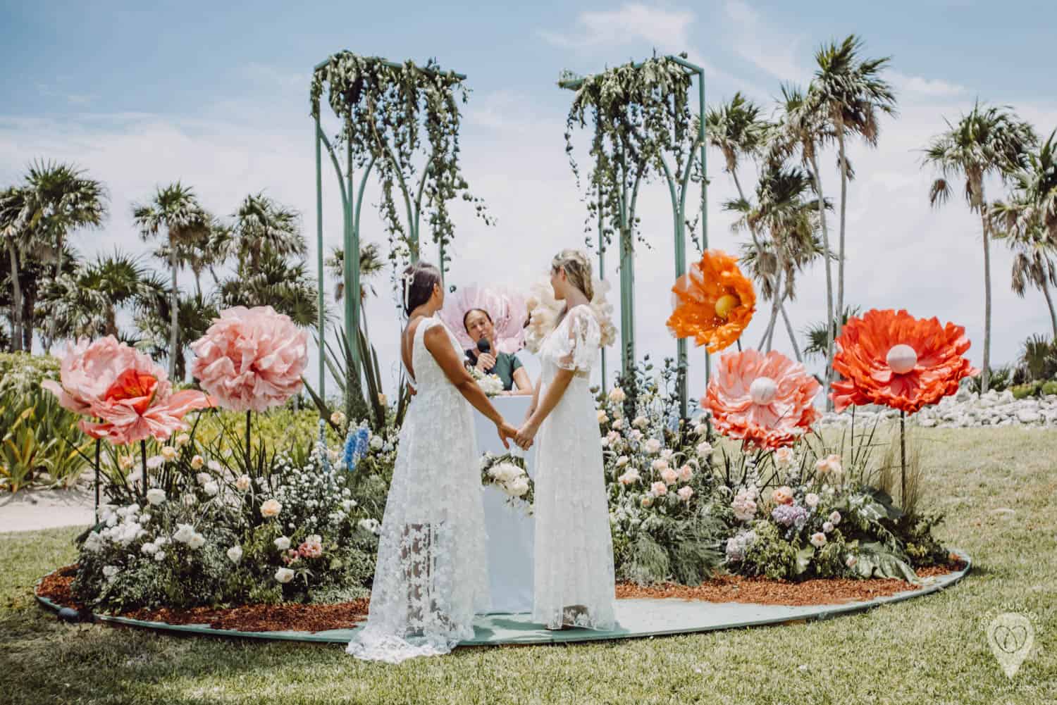 Two women in white dresses hold hands during an outdoor wedding ceremony, surrounded by large artificial flowers and greenery, with a celebrant officiating.
