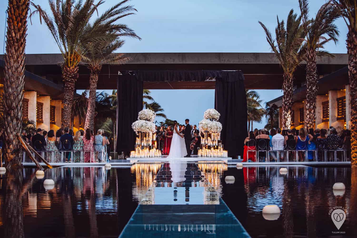 A couple stands at an outdoor wedding altar decorated with candles and white flowers, surrounded by guests seated on both sides of a reflective pool.