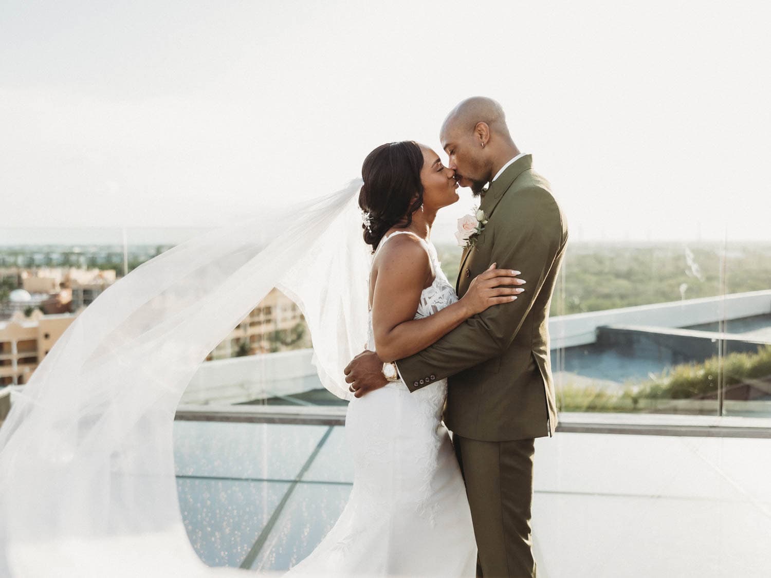 A bride and groom stand on a rooftop, embracing and kissing. The bride’s veil flows in the wind, and a scenic view is visible in the background.