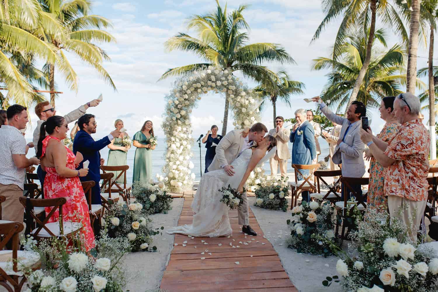 Couple kisses under a floral arch at an outdoor beach destination wedding, surrounded by cheering guests, palm trees, and white flowers.