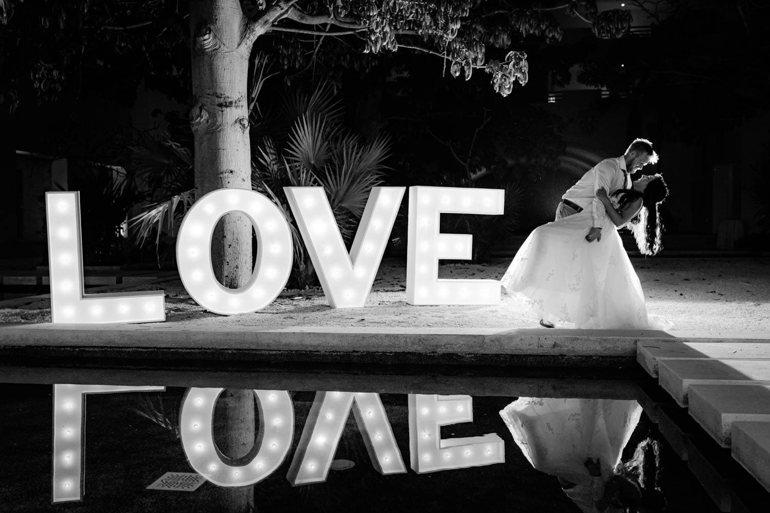 A couple poses in a romantic dip near large illuminated LOVE letters at night, with the scene reflected in nearby water.