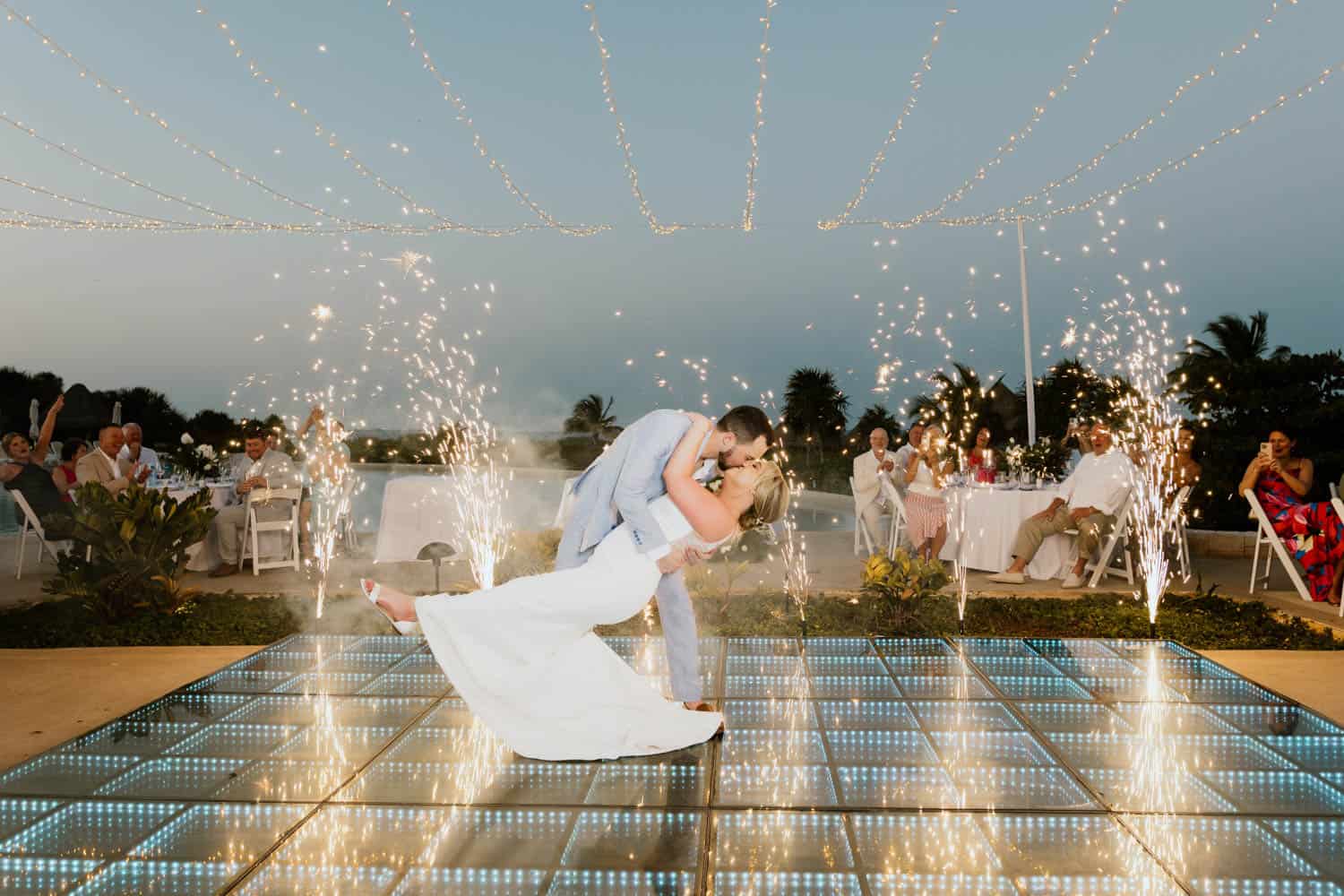 Bride and groom share a dip and kiss on a lit dance floor surrounded by sparklers at an outdoor evening destination wedding reception with guests in the background.