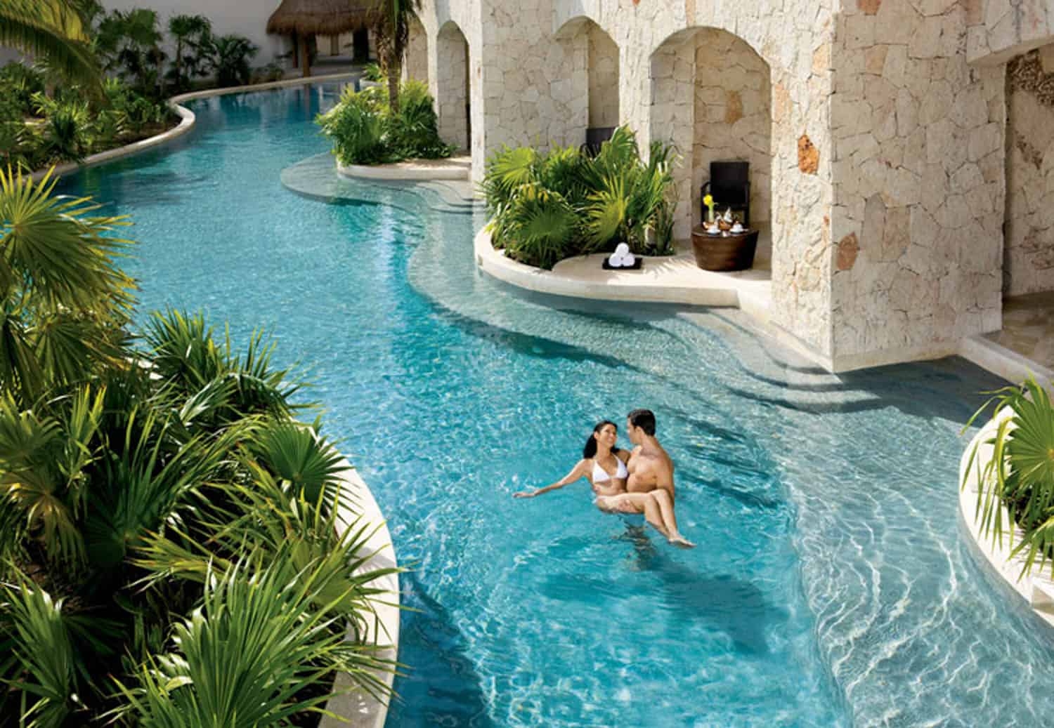 A couple stands in a clear, winding pool surrounded by lush greenery and stone walls, with lounge chairs and towels set up under shaded alcoves.