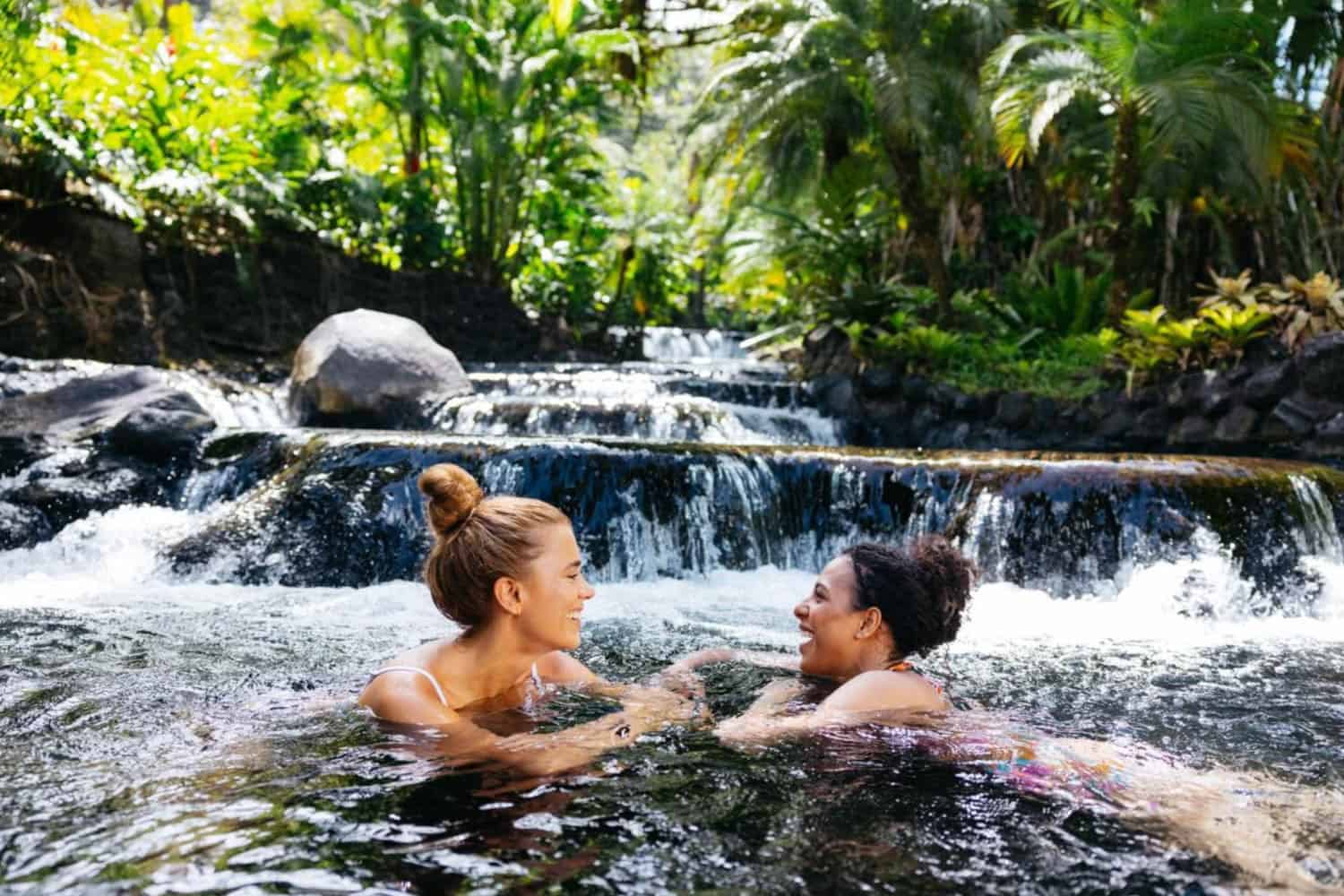 Two women are smiling and talking while swimming in a natural hot spring surrounded by lush greenery and a small waterfall in the background.