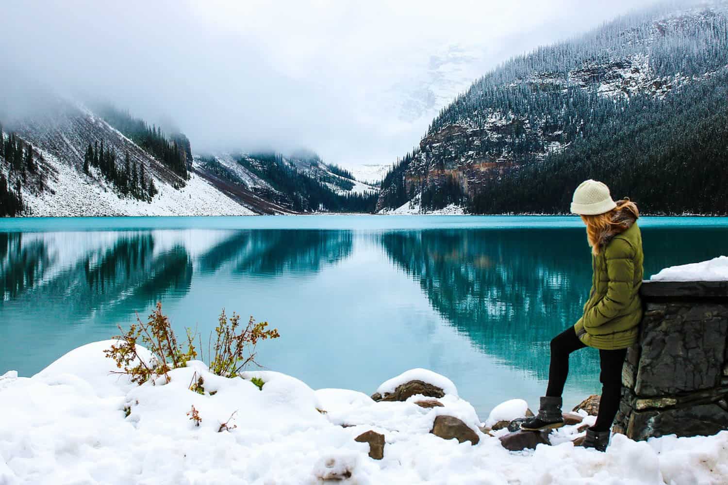A woman in winter clothing sits by a snowy lake, surrounded by snow-covered mountains and trees reflected in calm blue water under a cloudy sky.