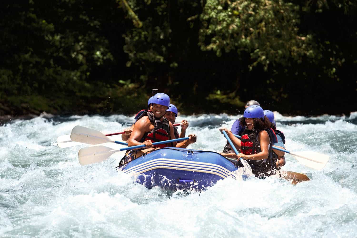 A group of people wearing helmets and life jackets paddle a blue inflatable raft through whitewater rapids on a river during an adventure travel vacation.