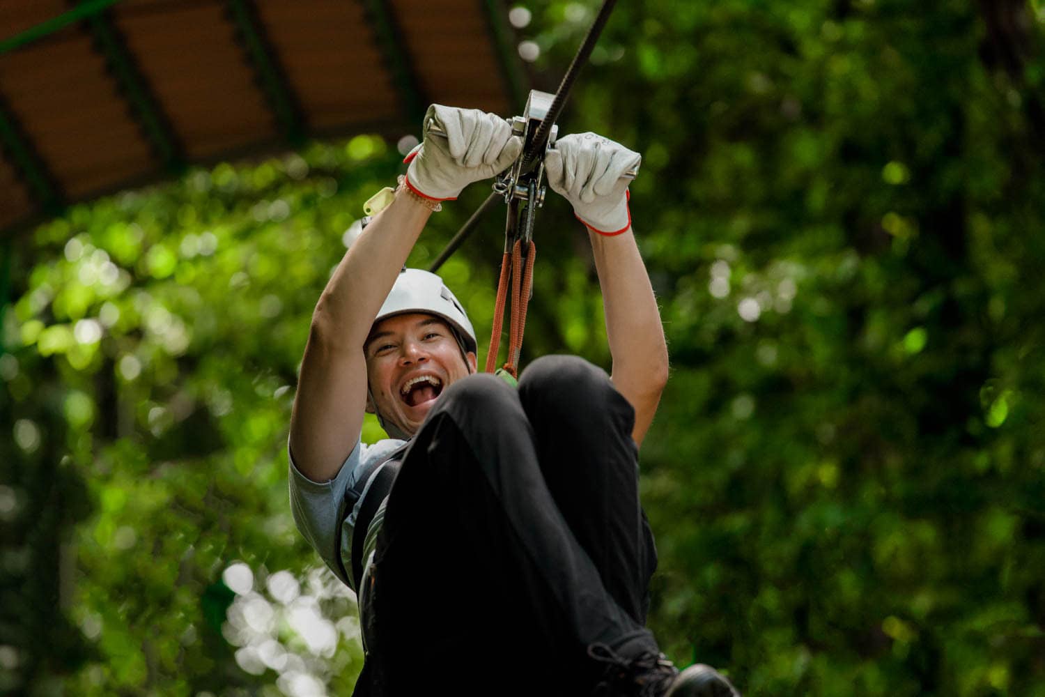 A man wearing a helmet and gloves rides a zip line through a green, forested area, smiling with excitement on an adventure travel trip.