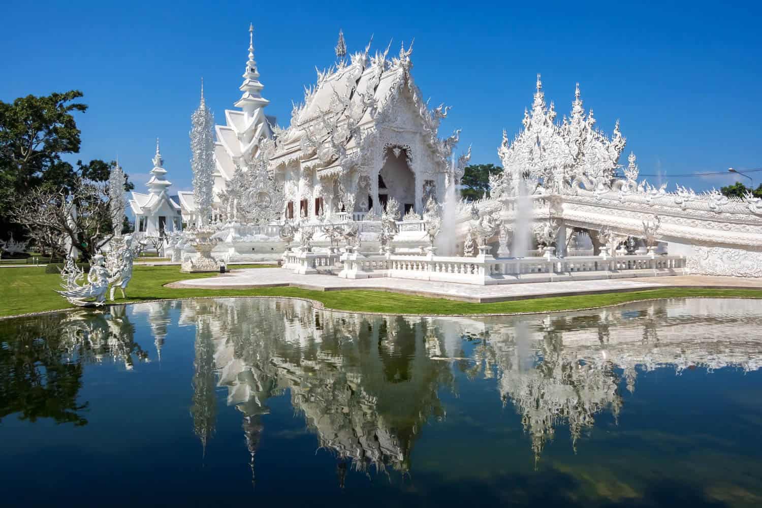 Wat Rong Khun, also known as the White Temple, in Chiang Rai, Thailand, stands beside a calm reflecting pool under a clear blue sky.