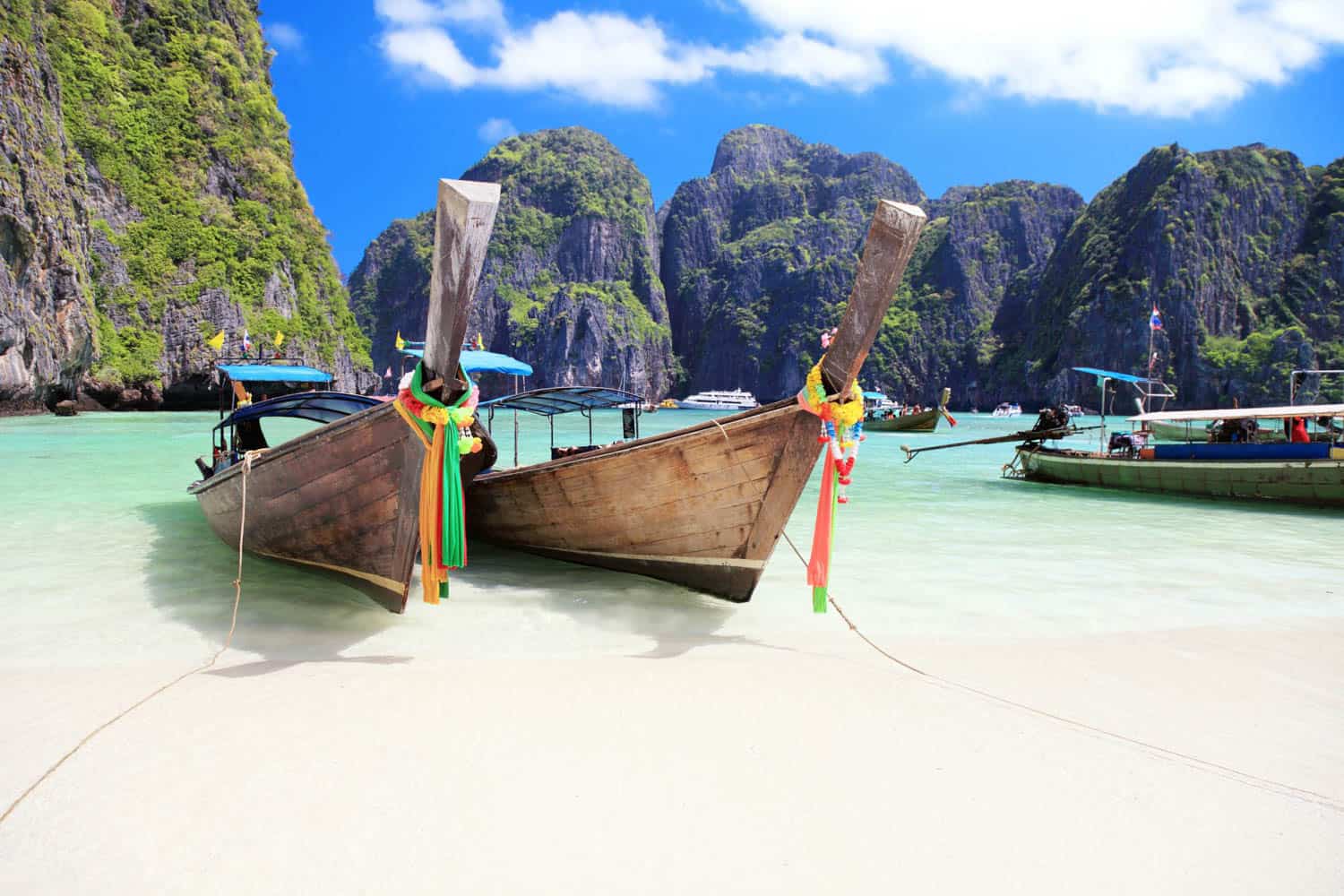 Two traditional wooden boats are anchored on a sandy beach with clear turquoise water and steep green cliffs in the background under a blue sky in Thailand.