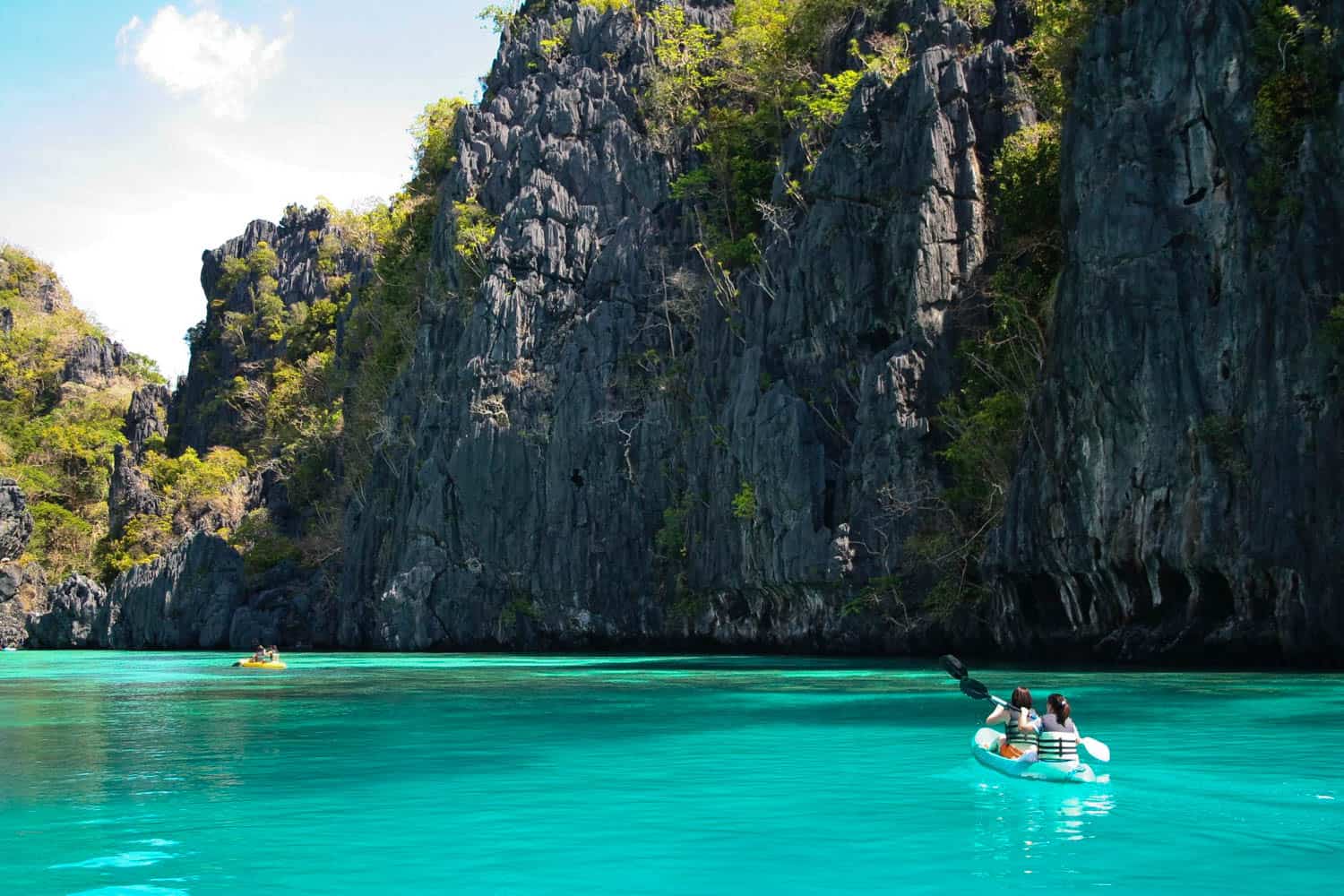 Two people kayak on clear turquoise water near tall, rocky cliffs covered with sparse greenery under a partly cloudy sky.