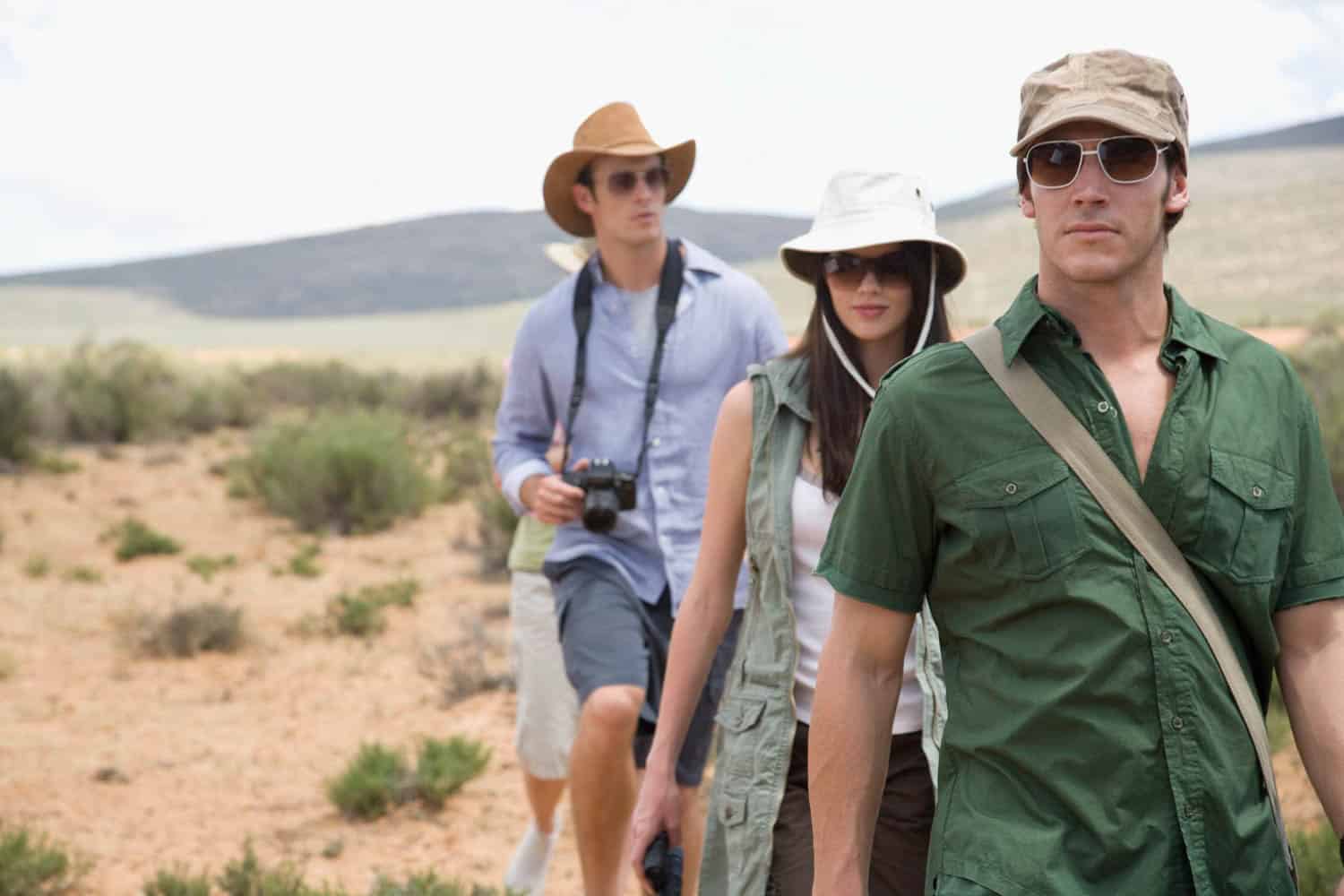 Four people wearing hats and sunglasses walk in a line through a desert landscape, while one holds a camera.