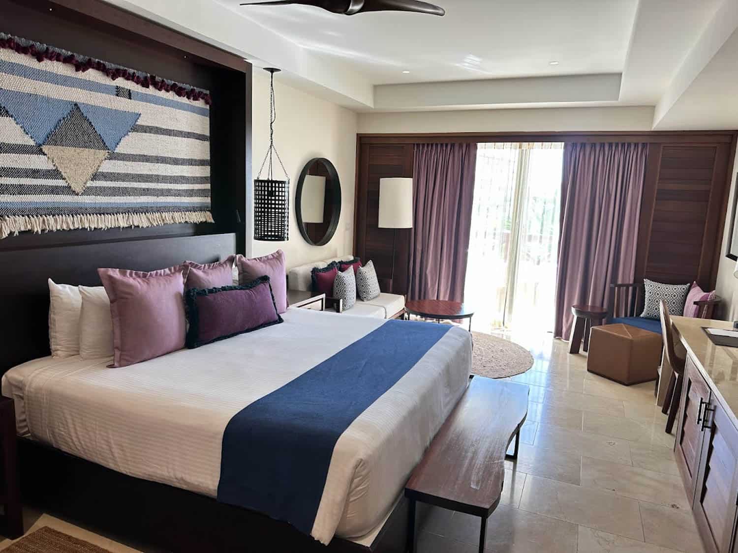JT-All-Inclusive-02 Modern hotel room with a large bed, purple and blue accents, seating area, wall art above the bed, and natural light coming from glass doors with purple curtains.