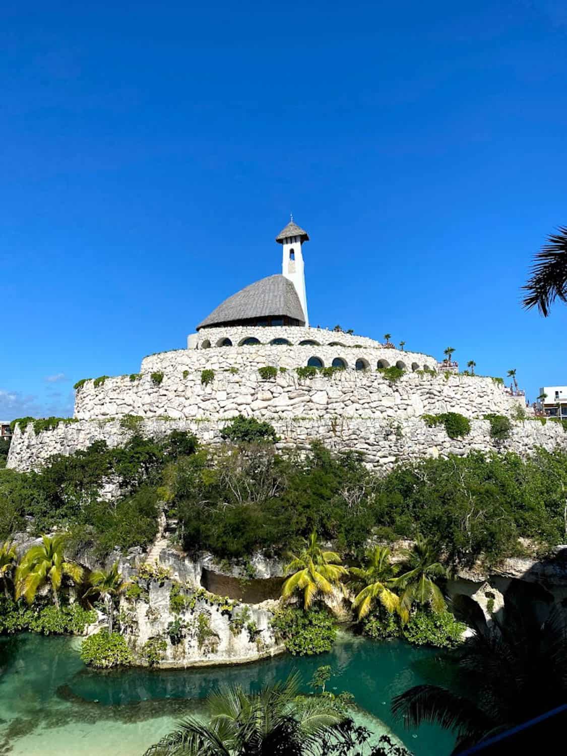 JT-All-Inclusive-03 A round stone building with a domed roof and small tower sits atop a rocky hill, surrounded by tropical plants and clear water under a blue sky.