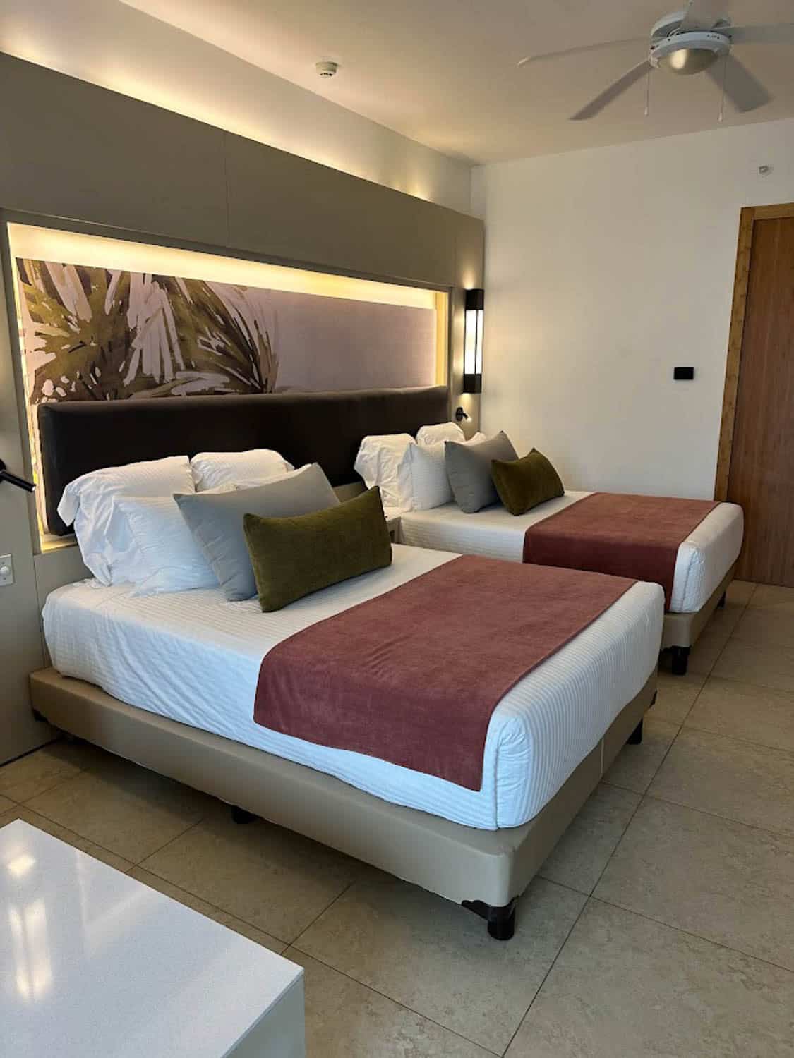 JT-All-Inclusive-04 An upscale hotel room with two double beds, white linens, mauve throws, green accent pillows, wall art, bedside lights, and a ceiling fan.