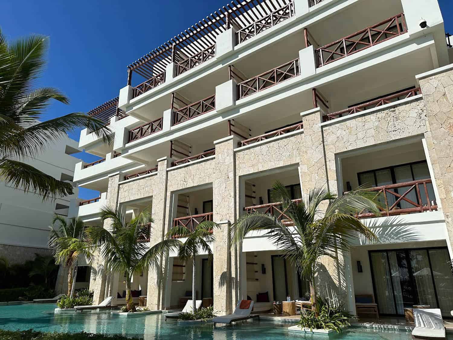 JT-All-Inclusive-06 Multi-story resort building with stone facade, balconies, and ground-floor pool access, surrounded by palm trees under a clear blue sky.