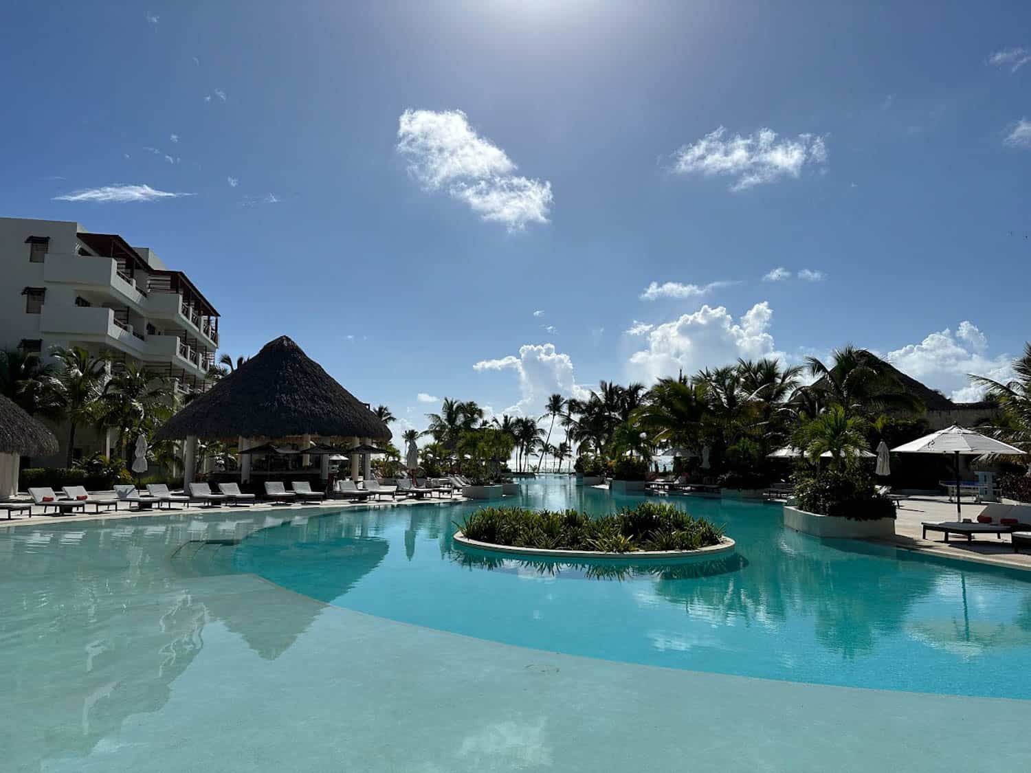 JT-All-Inclusive-07 Large outdoor pool at an all-inclusive resort with lounge chairs, thatched-roof bar, palm trees, and clear blue sky under bright sunlight.