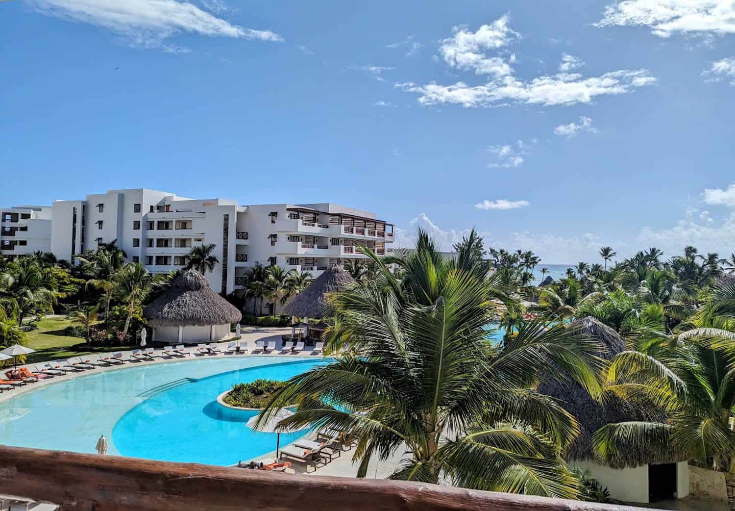 JT-All-Inclusive-08 A resort with a large outdoor pool, lounge chairs, palm trees, and multi-story white buildings under a blue sky with scattered clouds.