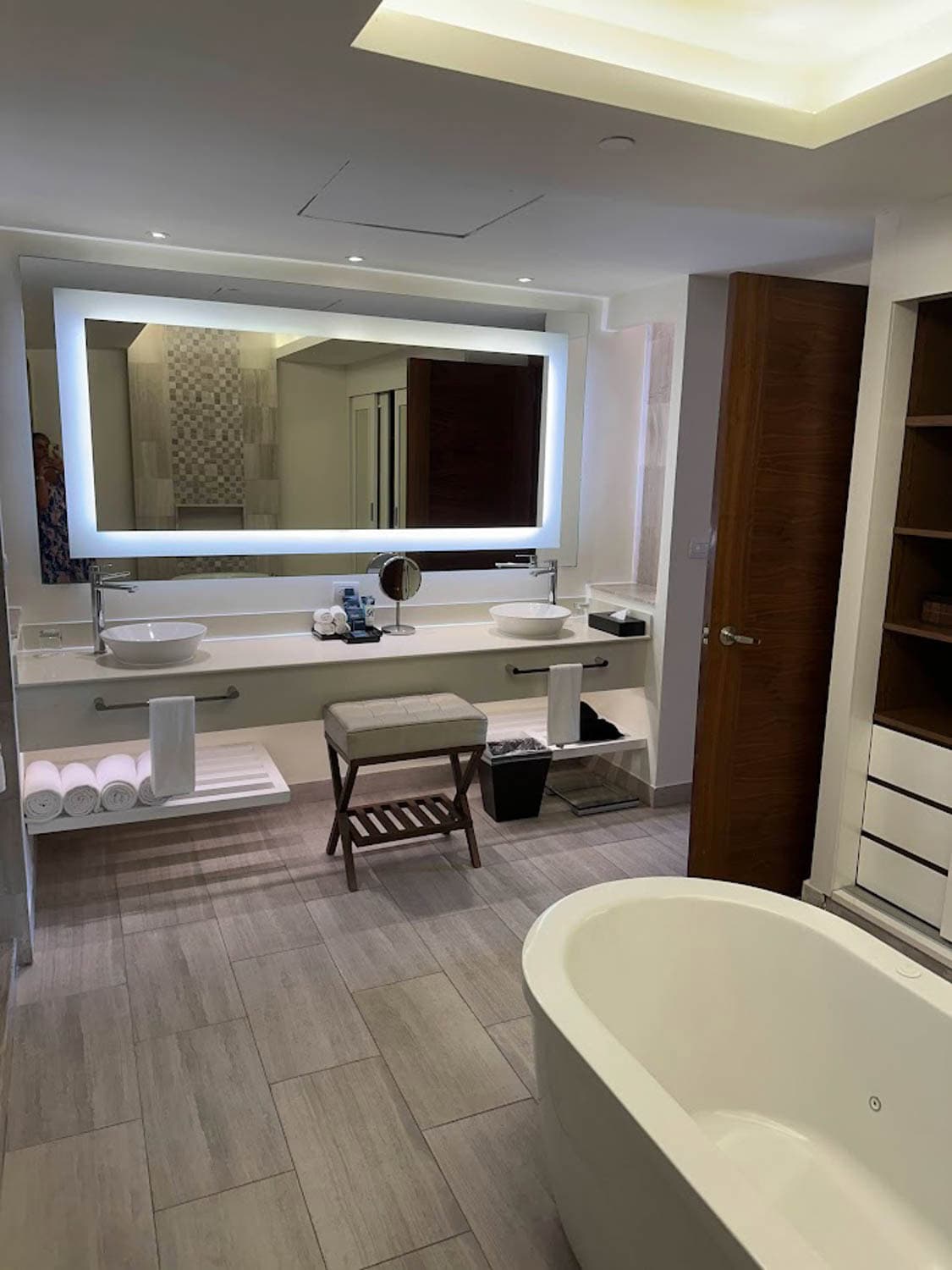 JT-All-Inclusive-09 Modern bathroom with a large illuminated mirror, double sinks, a freestanding tub, wooden accents, and neatly rolled towels on a shelf beneath the countertop.