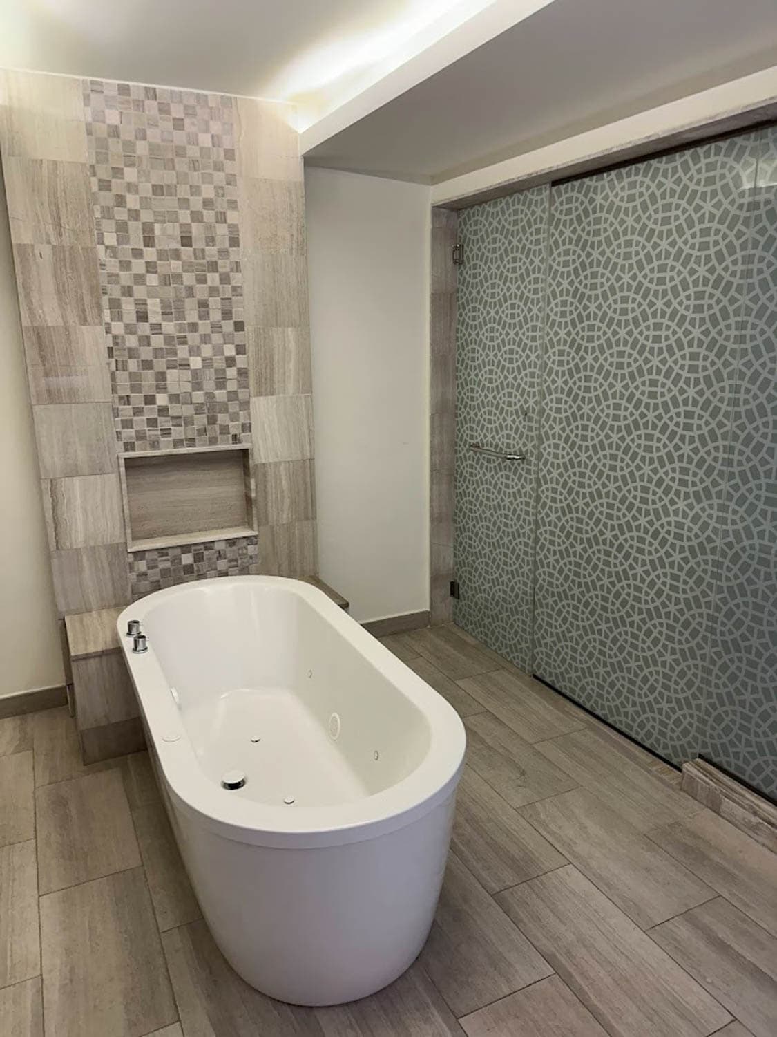 JT-All-Inclusive-10 Modern bathroom with a freestanding white bathtub, patterned tile accent wall, and frosted glass shower doors with geometric designs.