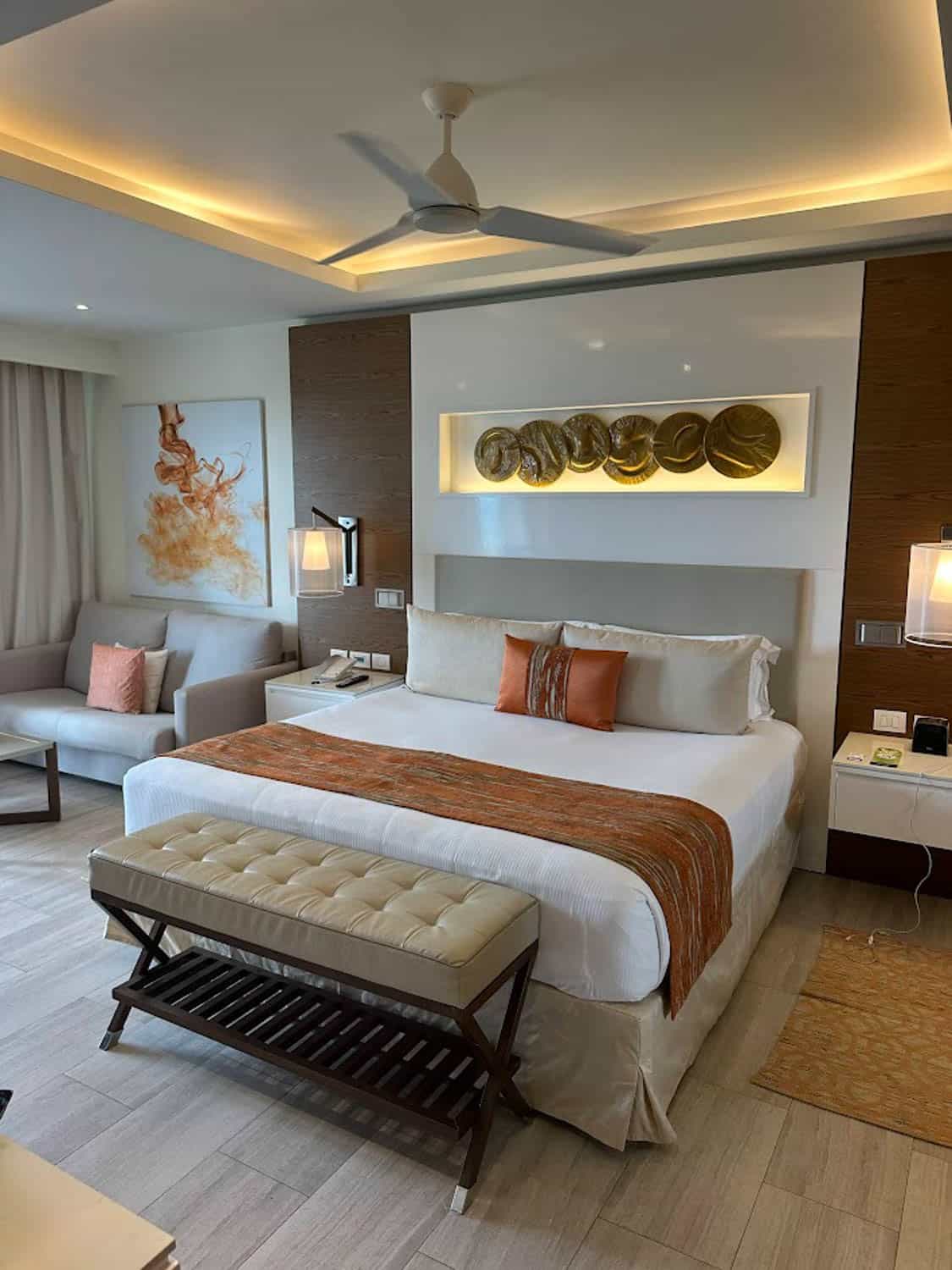JT-All-Inclusive-11 A modern hotel room with a large bed, orange accents, a tufted bench, wall art, bedside tables, a sofa, and ceiling fan under soft lighting.