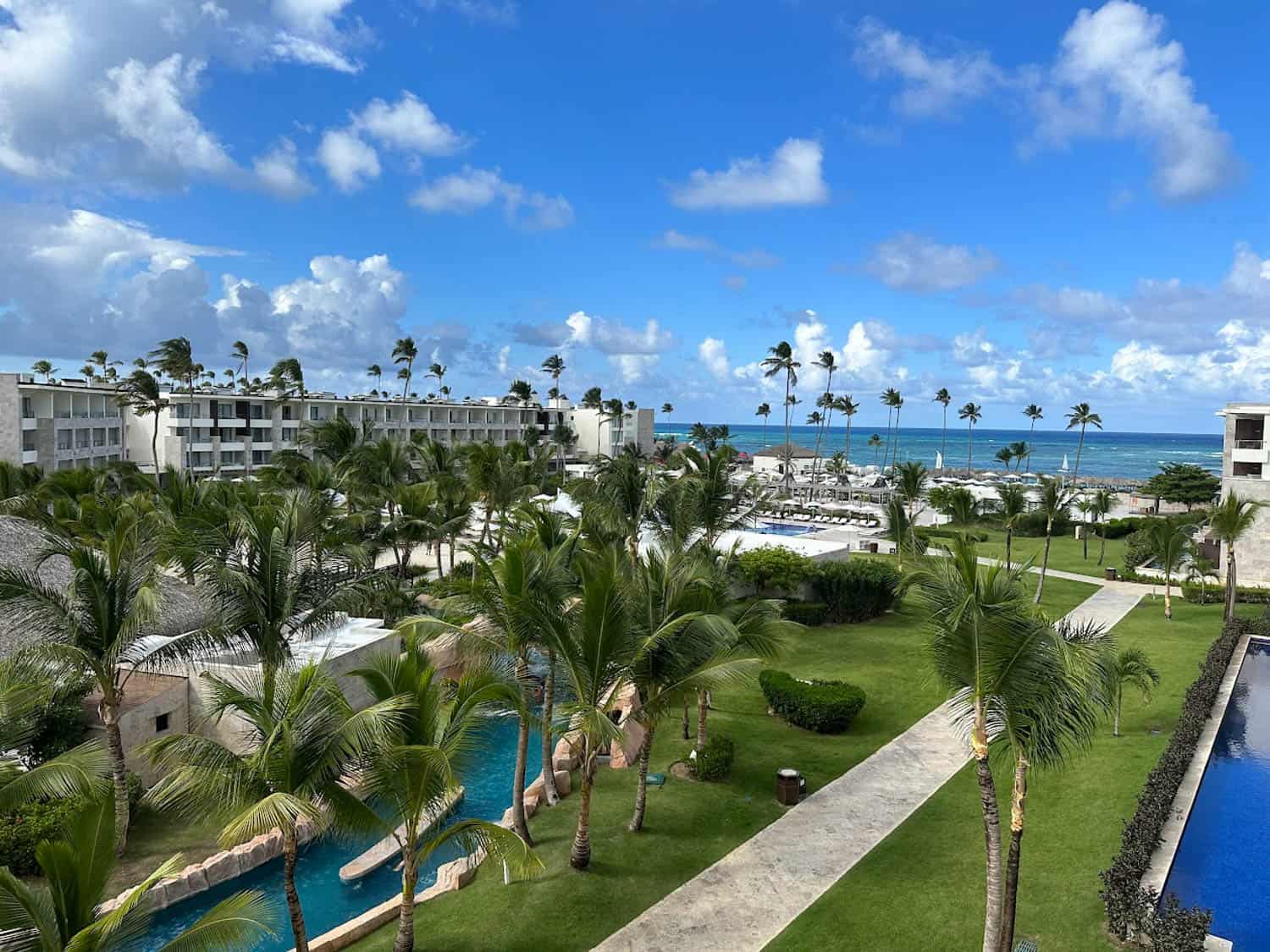 JT-All-Inclusive-12 View of an all-inclusive resort with palm trees, a swimming pool, green lawns, and buildings under a partly cloudy blue sky. The ocean is in the background.