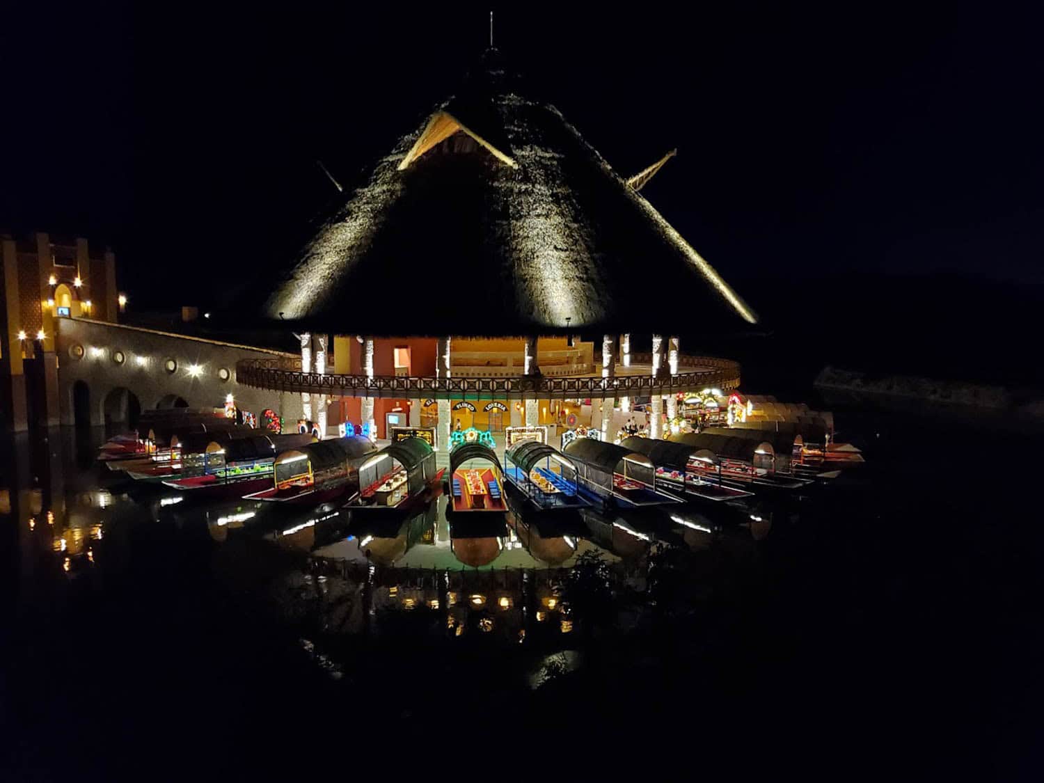 JT-All-Inclusive-15 Thatched-roof building at night with bright lights, surrounded by water. Several colorful boats are docked in front, reflecting on the calm surface.