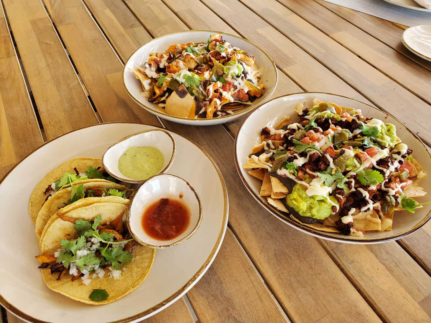 JT-All-Inclusive-16 3 tacos with cilantro and onions, and two sauces, alongside two bowls of loaded nachos with beans, guacamole, pico de gallo, and sour cream on a wooden table.
