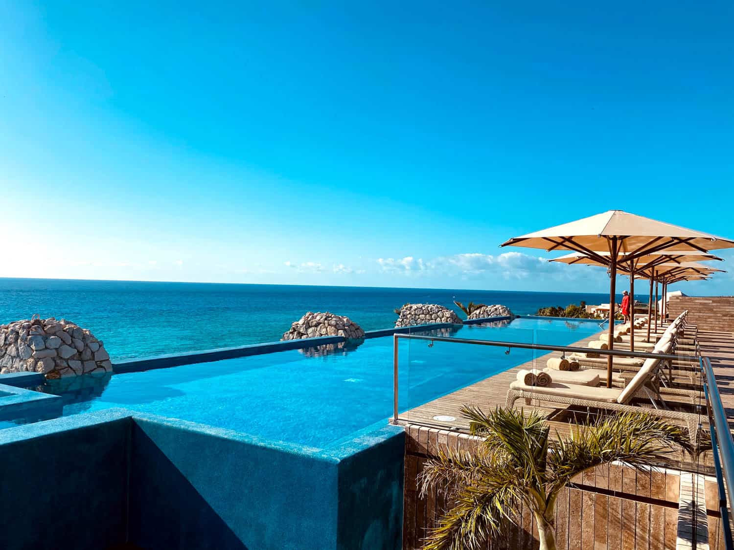 JT-All-Inclusive-17 Infinity pool overlooking the ocean with deck chairs, umbrellas, and a clear blue sky at an all-inclusive resort.