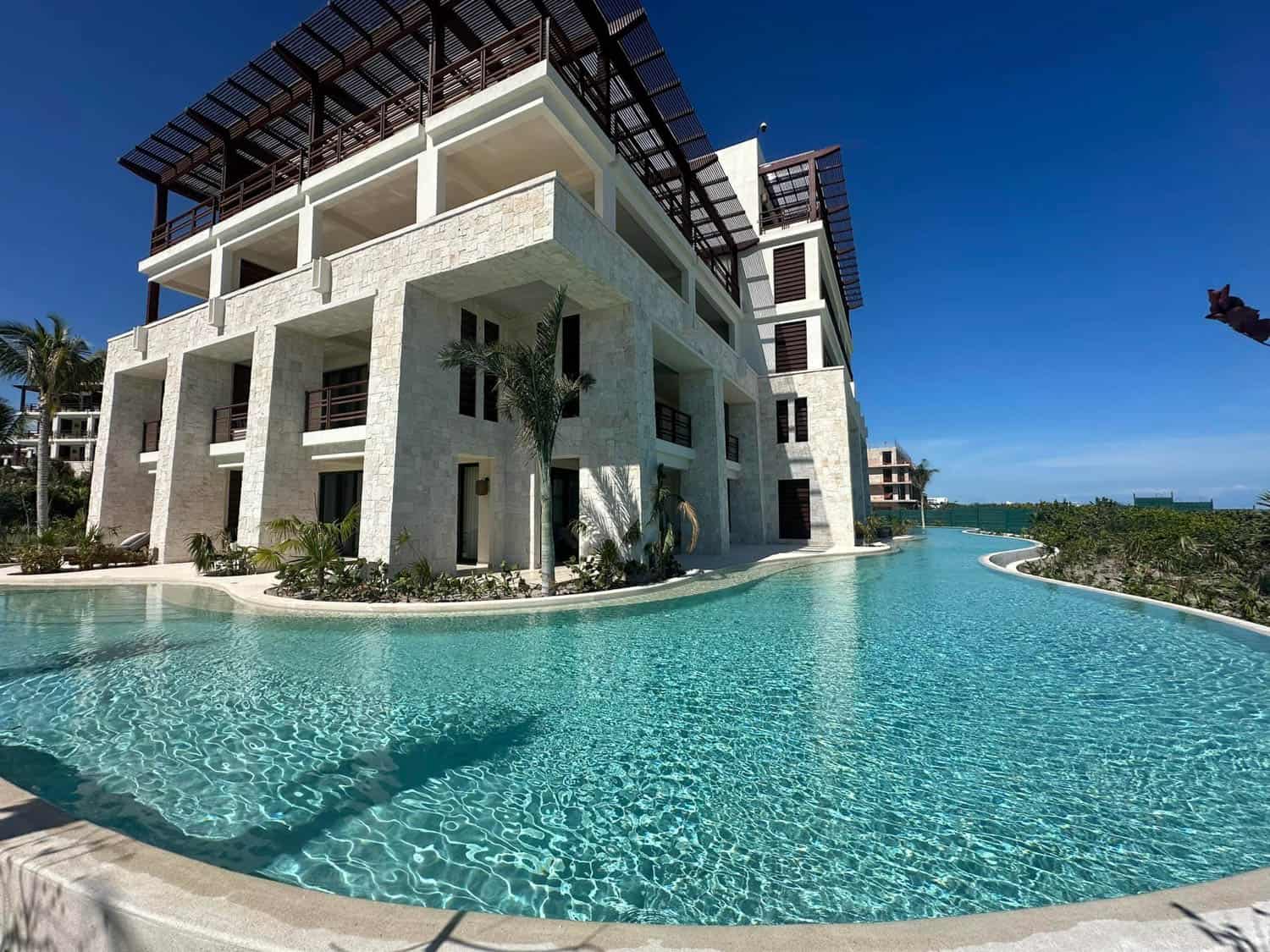Modern Caribbean resort building with large balconies and white stone exterior surrounded by a clear, winding swimming pool under a bright blue sky.