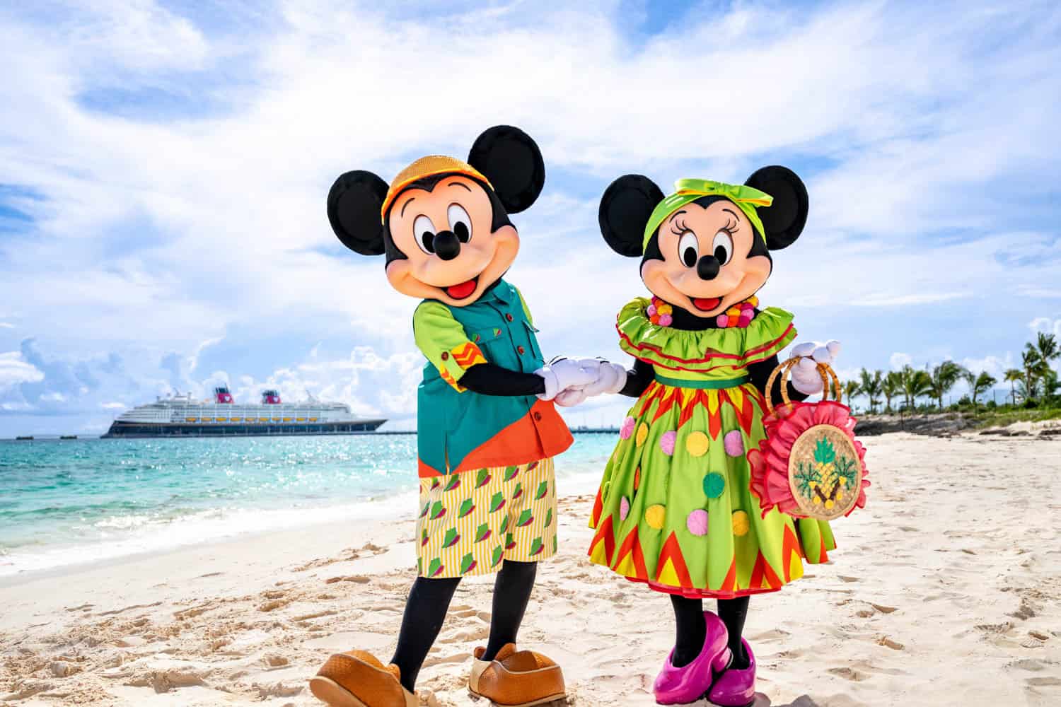 JT-Disney-01 Mickey Mouse and Minnie Mouse in colorful tropical outfits standing together on a sandy beach with a cruise ship and palm trees in the background.