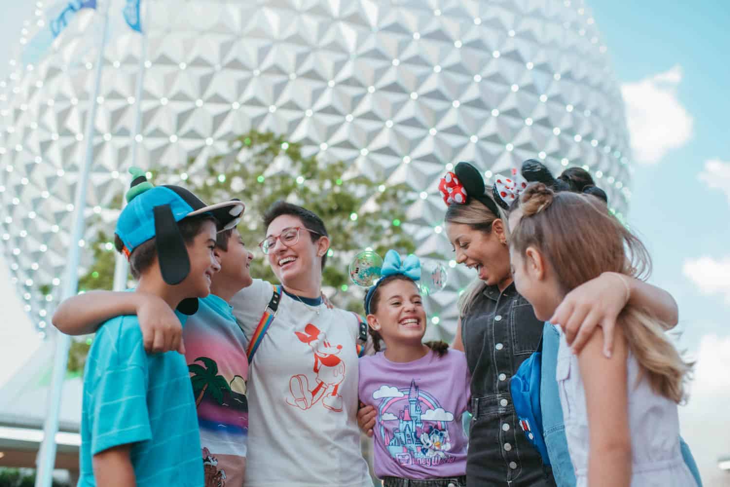 JT-Disney-02 A group of six people, including children and adults, stand together smiling in front of the Epcot sphere at Disneyland, wearing colorful clothes and themed headbands.