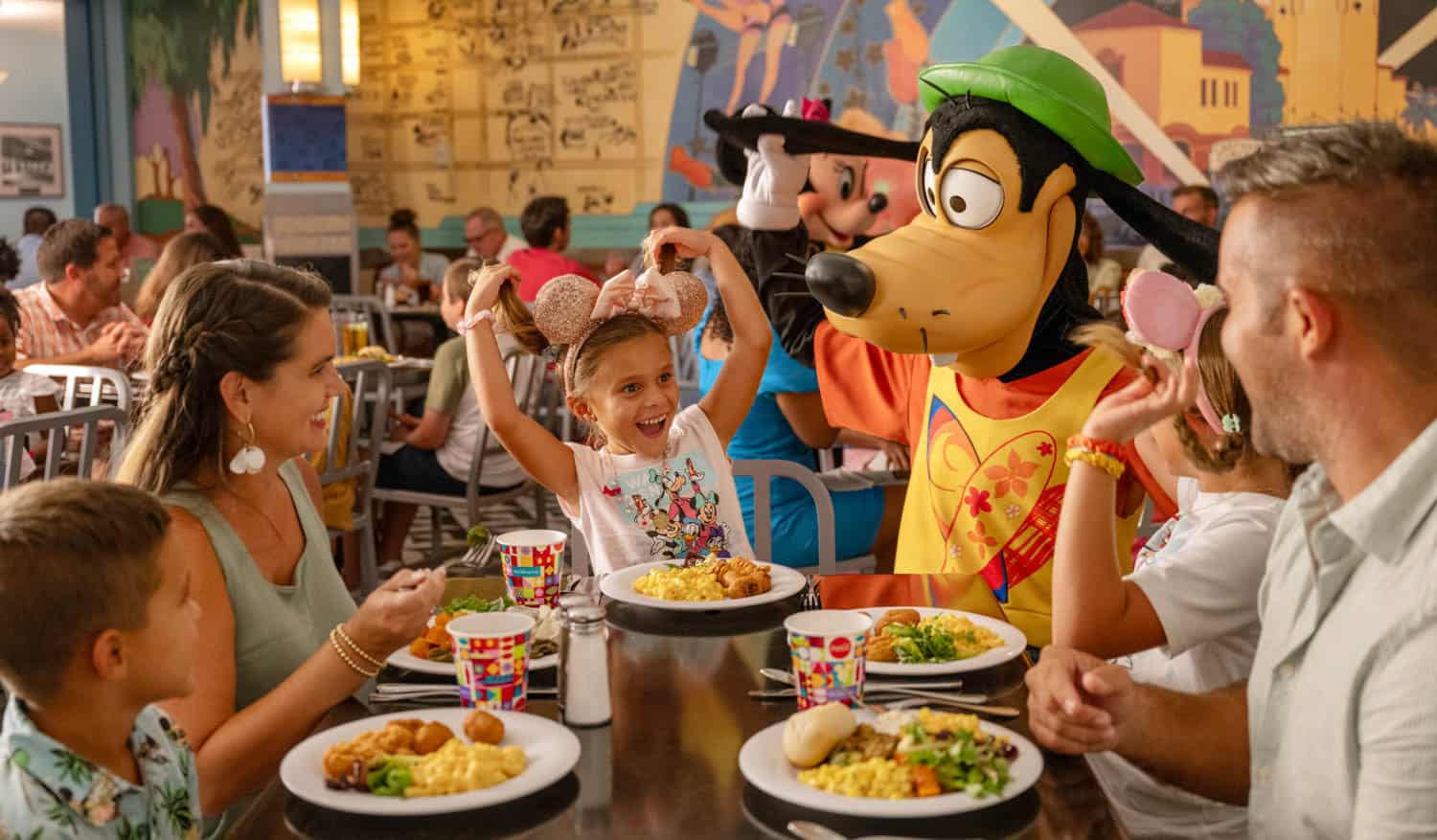 JT-Disney-06 A family sits at a restaurant table while on a Disney vacation, as a person in a Goofy costume interacts with a smiling child wearing Minnie Mouse ears.