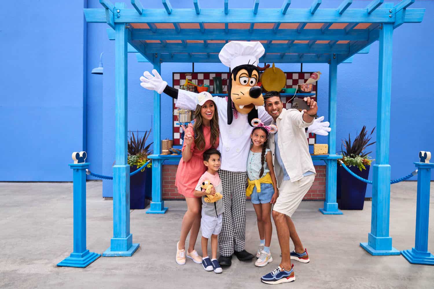 JT-Disney-08 A family of four on their Disney vacation, poses and smiles with a person in a Goofy costume dressed as a chef, standing under a blue pergola.