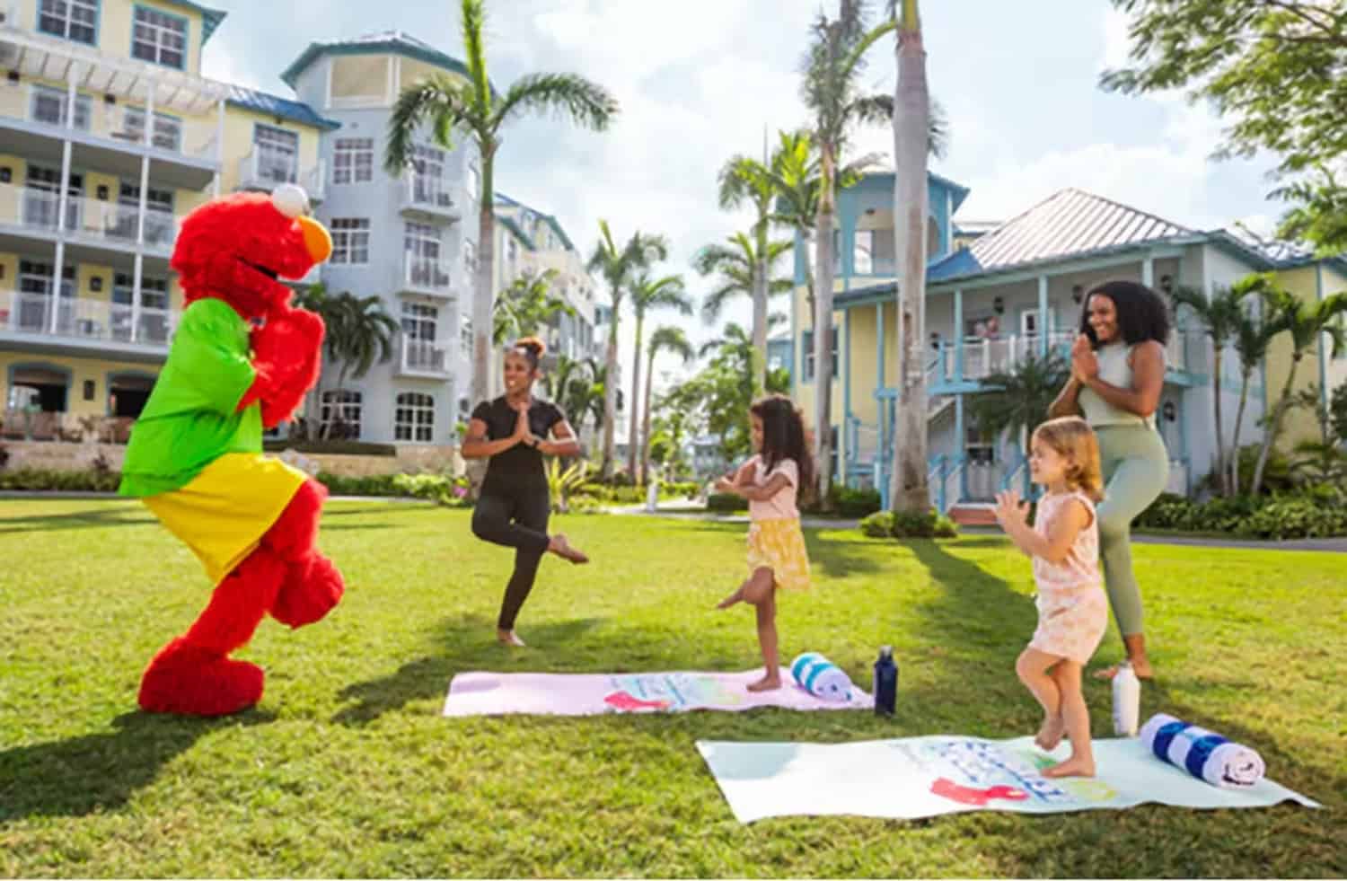 JT-Family-Travel-01 A person in an Elmo costume leads a family yoga session with two adults and two children on mats outside near palm trees and resort buildings.