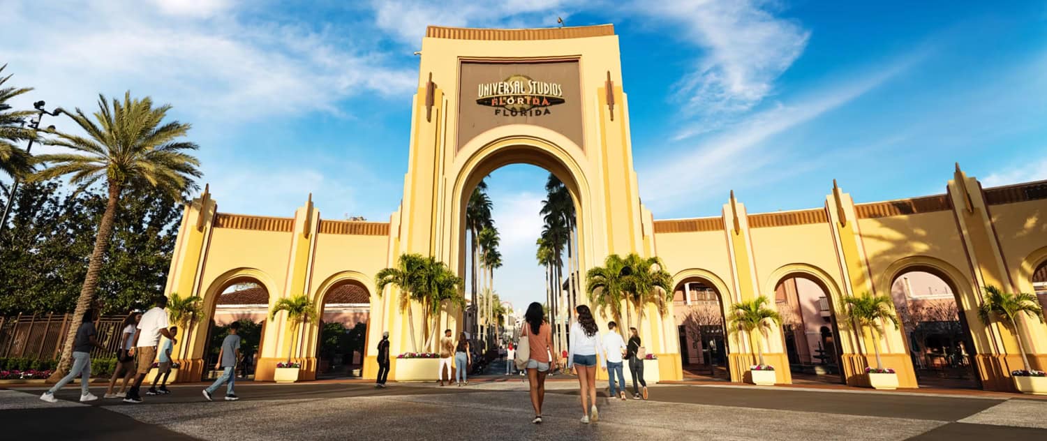 Screenshot People walk near the main entrance arch of Universal Studios Florida, surrounded by palm trees and bright yellow buildings under a sunny blue sky.