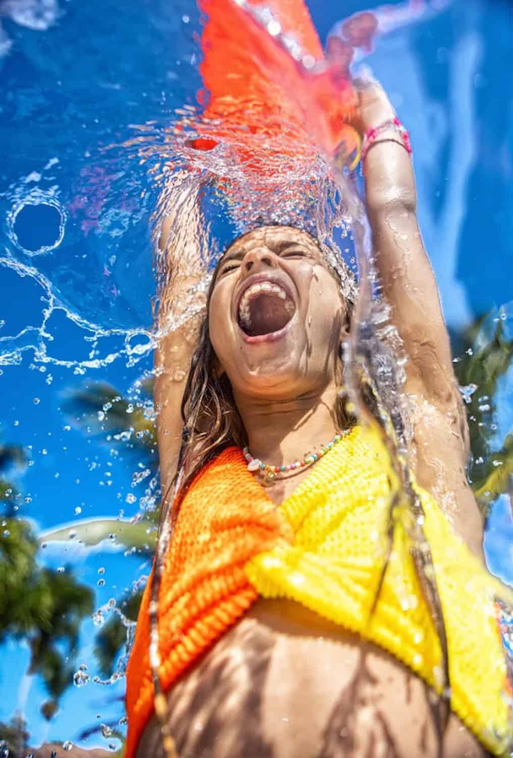 JT-Family-Travel-04 Child wearing an orange and yellow top smiles with arms raised under splashing water on a sunny day, blue sky and palm trees in the background.