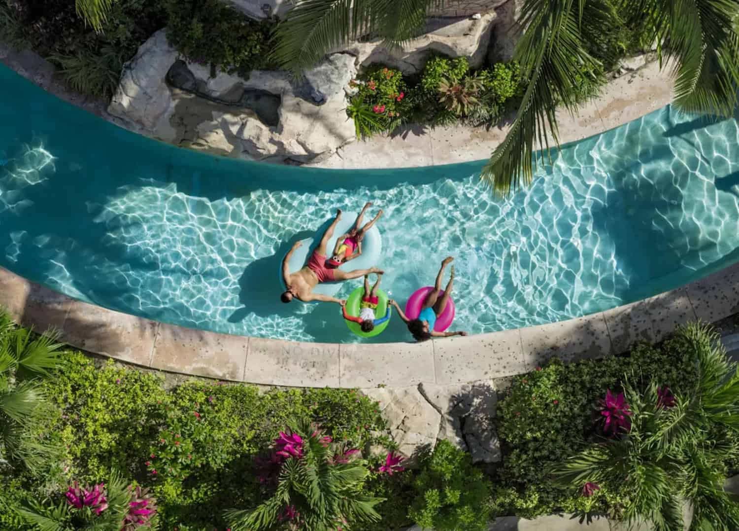JT-Family-Travel-05 Aerial view of three people floating on inflatable tubes in a winding outdoor pool surrounded by tropical plants and palm trees.
