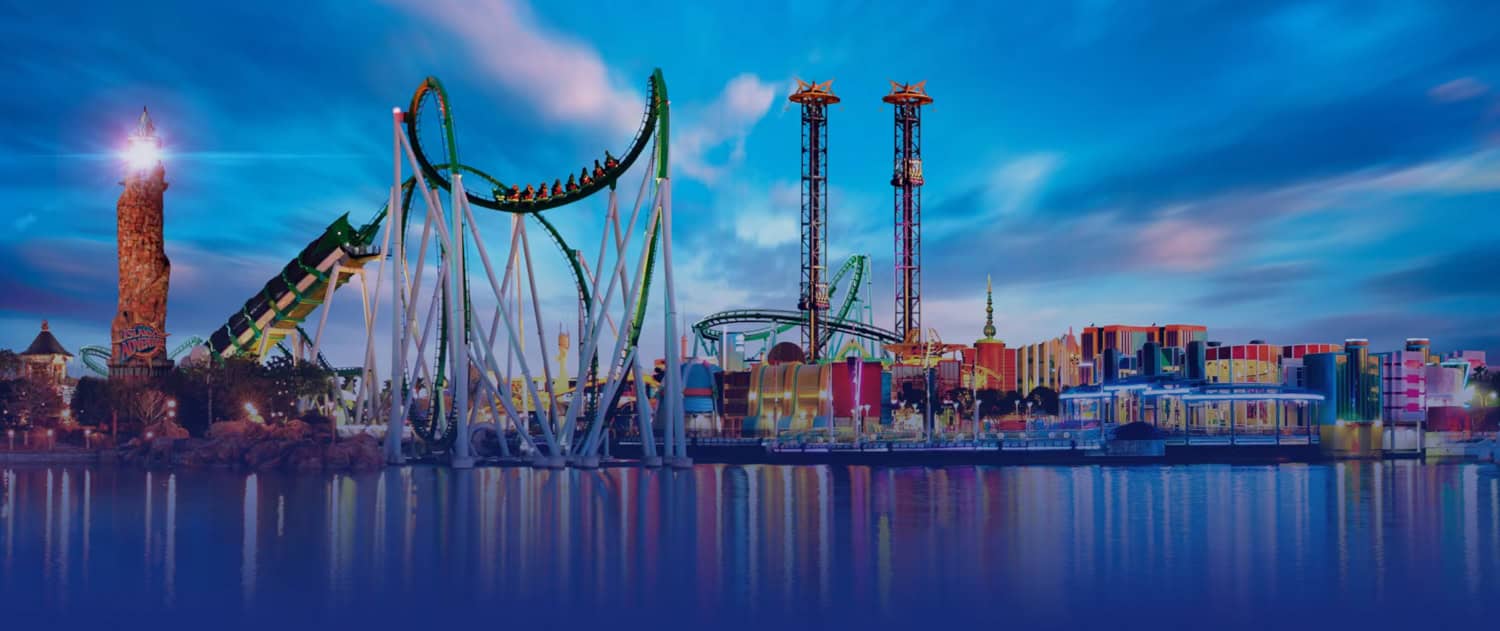 Screenshot A theme park ideal for family travel at dusk featuring roller coasters, tall ride towers, colorful buildings, and water in the foreground under a blue sky.