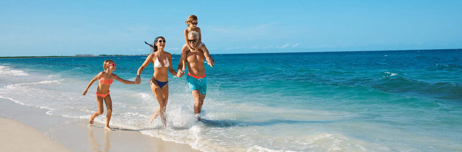 JT-Family-Travel-08 A family of four in swimsuits walks along the shoreline on a sunny beach, with waves splashing around them and the ocean in the background.
