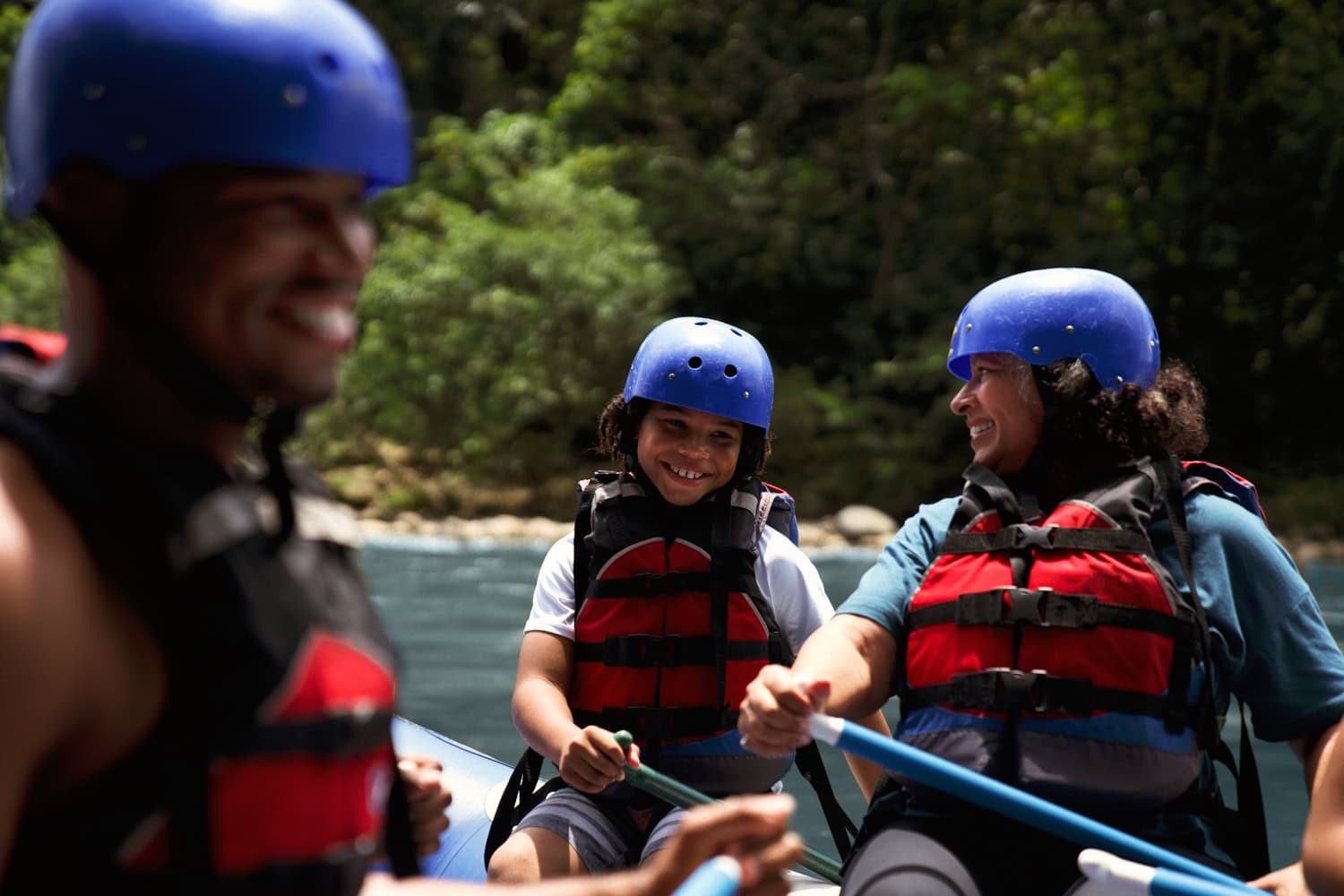 JT-Family-Travel-10 Three people wearing life jackets and helmets are white-water rafting on a river, surrounded by trees.