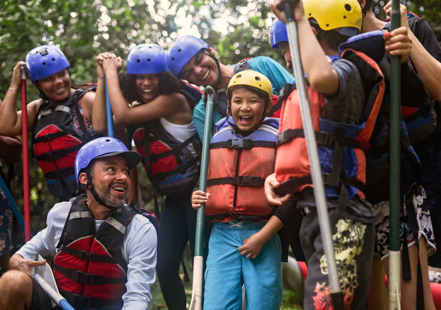 JT-Family-Travel-11 A group of people wearing helmets and life jackets stand together outdoors, holding paddles and smiling.